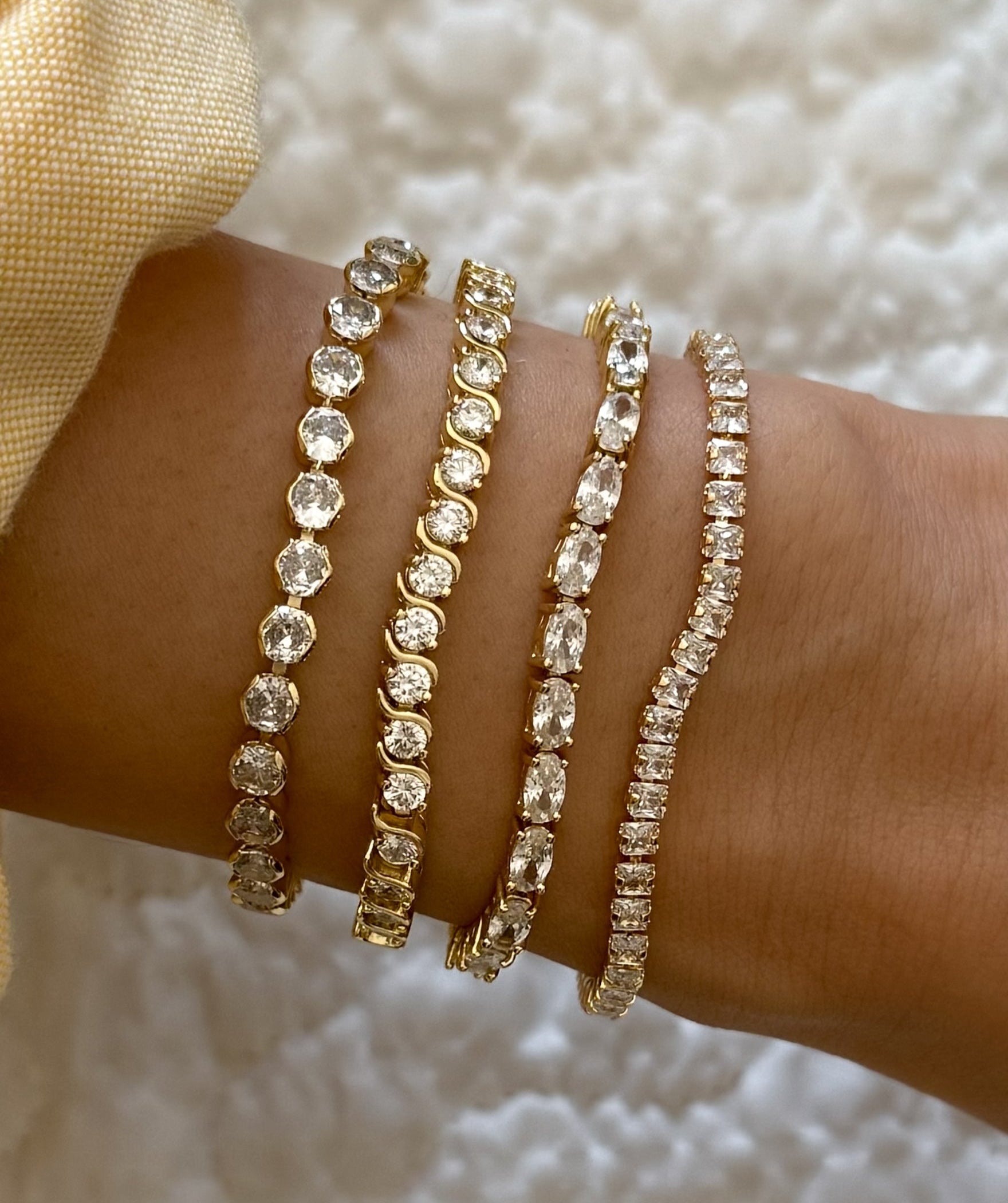Close-up of a wrist wearing multiple gold and crystal tennis bracelets on a textured white background.