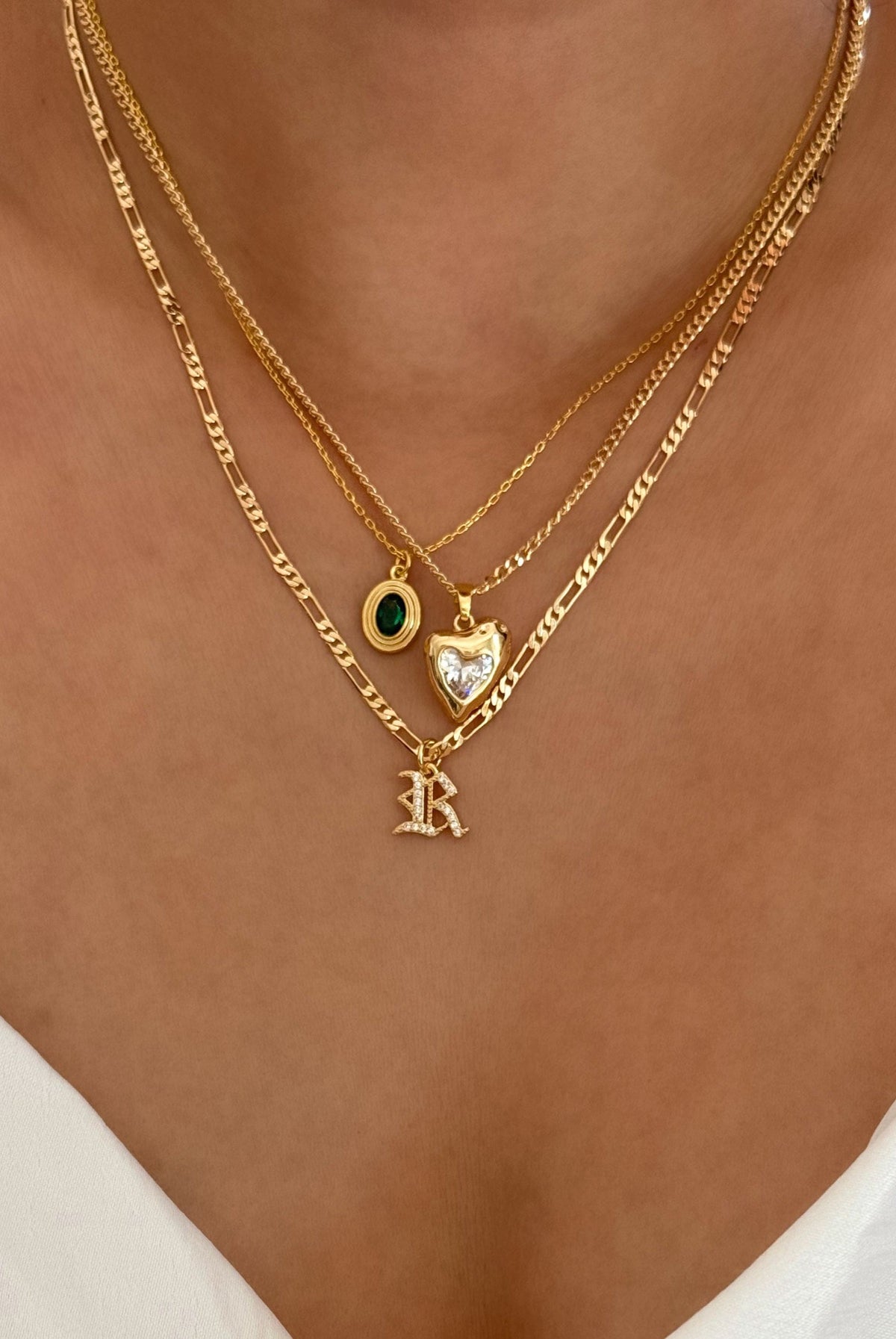 Gold necklaces with heart and letter 'R' pendant on a person wearing a white top.