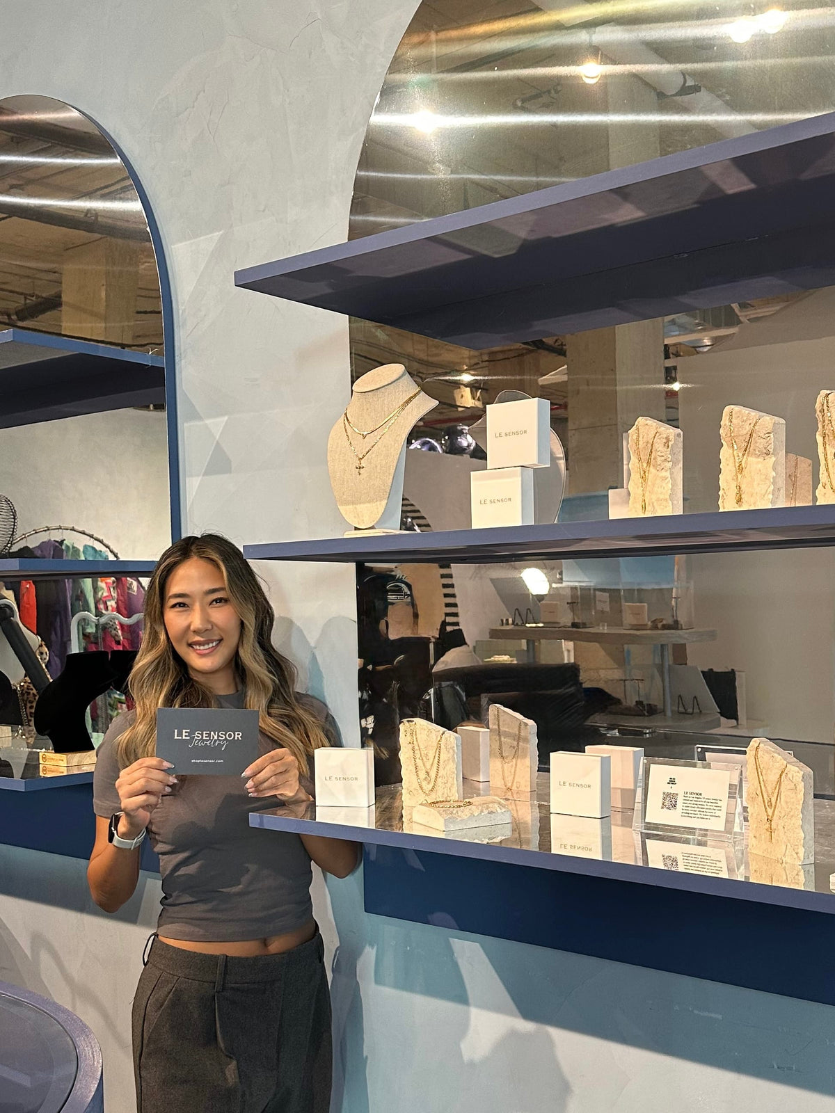 A woman stands in a jewelry store display area holding a card that reads 'LE SENIOR'. Shelves behind her display necklaces on busts and boxes, with blue shelving and a light gray wall.