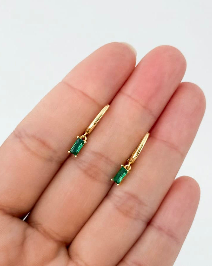Gold earrings with green gemstones held between fingers on a white background