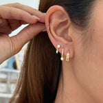 A close-up of a models ear wearing a stack of gold earrings, including a small baguette row huggies, a dangling pearl-like stud, and a vertical bar earring, with her hand adjusting the top earring.