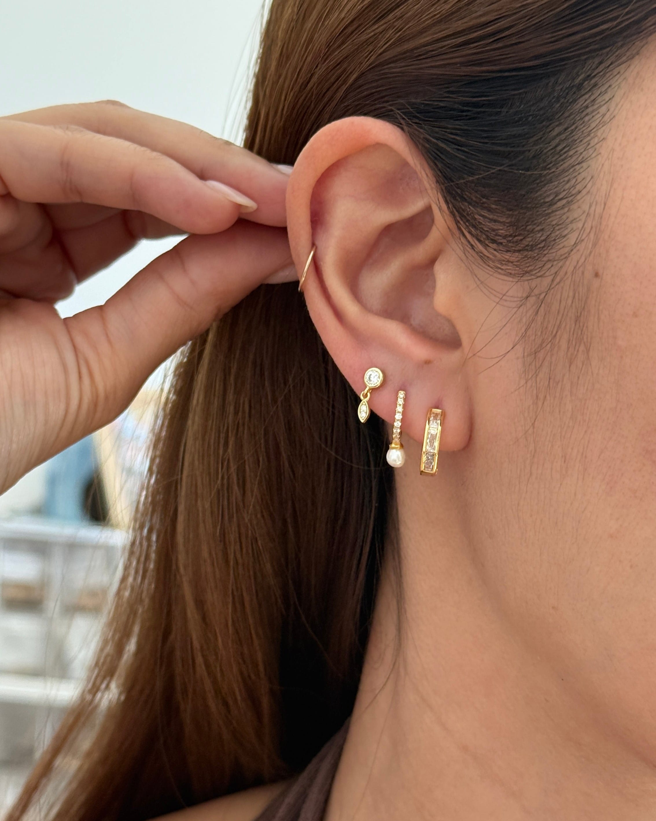 A close-up of a models ear wearing a stack of gold earrings, including a small baguette row huggies, a dangling pearl-like stud, and a vertical bar earring, with her hand adjusting the top earring.