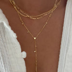 A close-up of a person wearing a layered gold necklace set over a white textured top. The necklace features multiple chain styles, including a curb chain, a figure-eight chain, and a delicate lariat Y-shaped chain with small star and dot charms.
