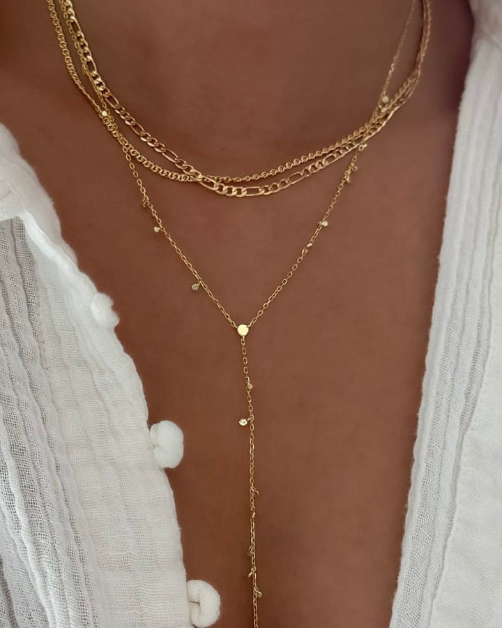 A close-up of a person wearing a layered gold necklace set over a white textured top. The necklace features multiple chain styles, including a curb chain, a figure-eight chain, and a delicate lariat Y-shaped chain with small star and dot charms.