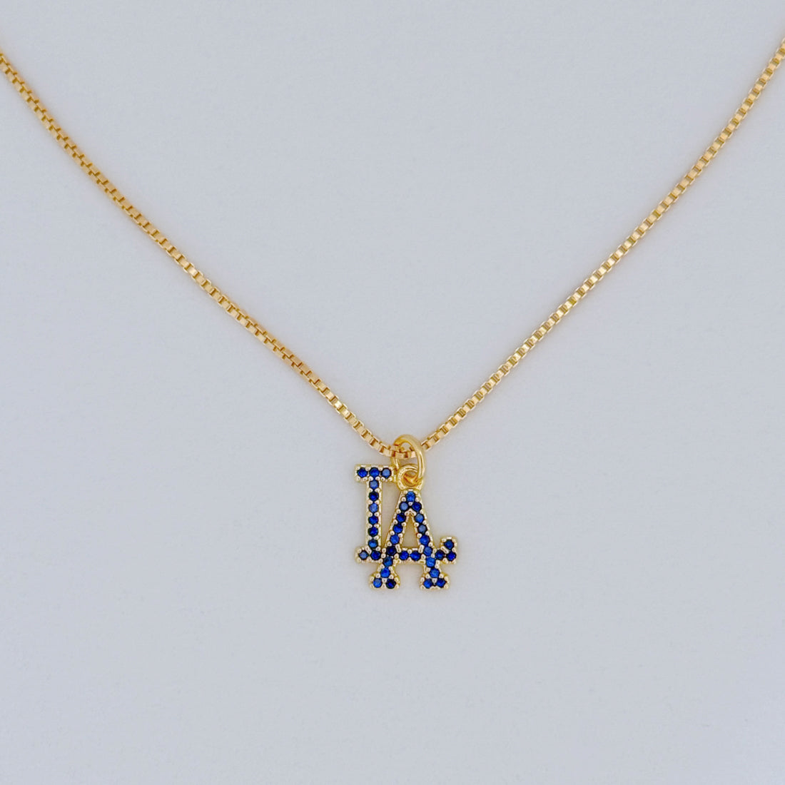 Gold necklace with a blue 'LA' pendant on a light gray background