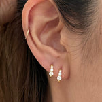 A close-up of a person's ear wearing a pair of small gold huggie hoop earrings with clear gemstones, along with a thin gold ear cuff.