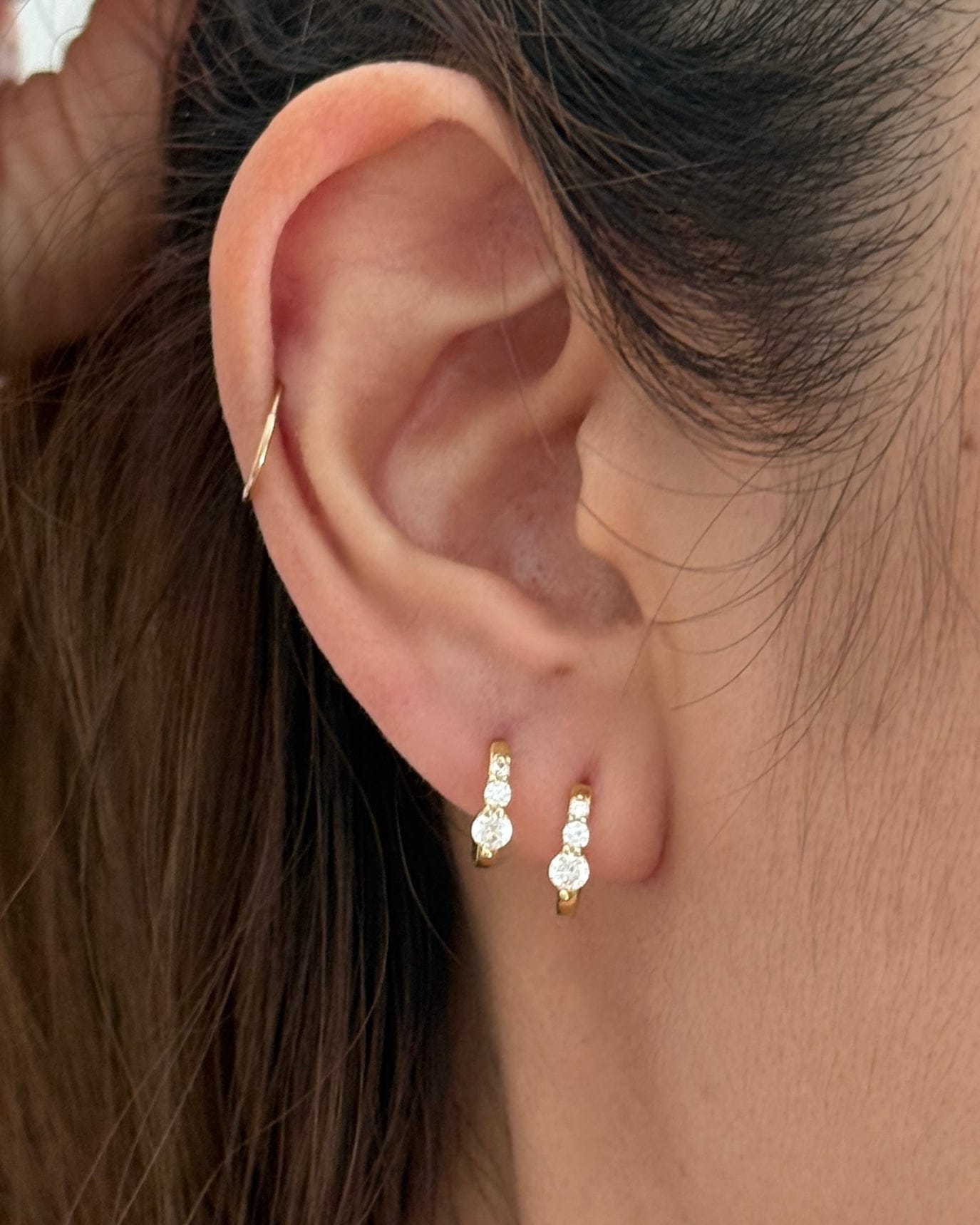 A close-up of a person's ear wearing a pair of small gold huggie hoop earrings with clear gemstones, along with a thin gold ear cuff.