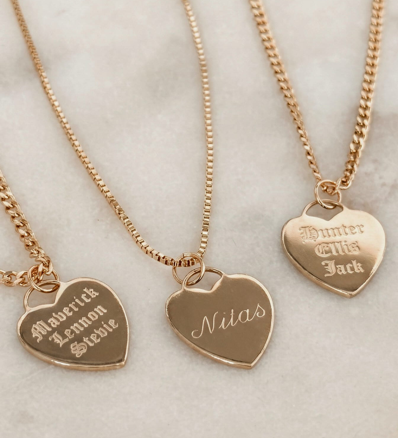 Three personalized gold heart pendant necklaces on matching gold chains, displayed on a light marble surface. The heart pendants are engraved with names: 'Maurice & Lemon Sviee', 'Nitas', and 'Hunter & This Jack'.