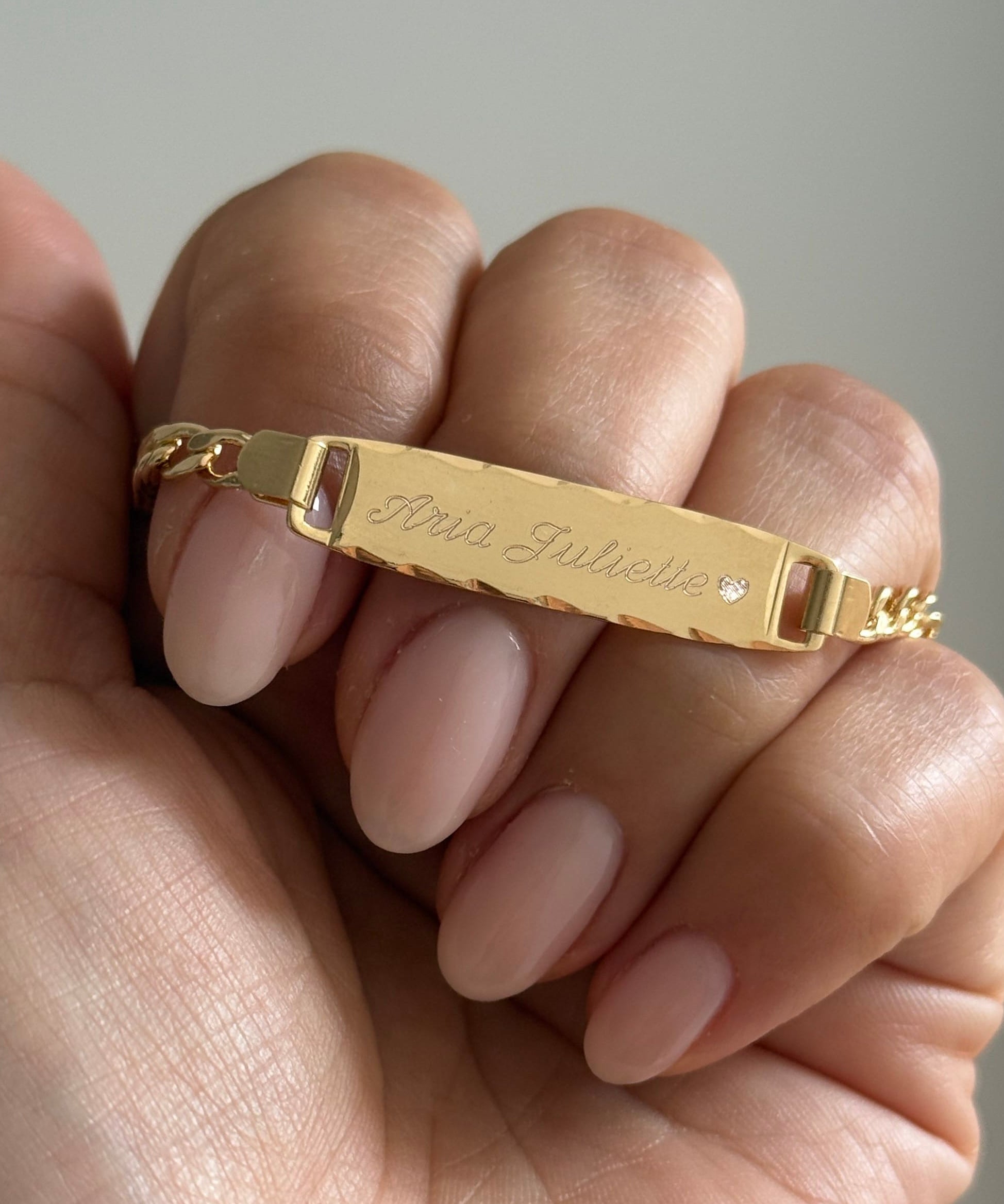 A close-up of a hand holding a gold ID bracelet engraved with the name 'Anna Juliebie' and a small clear stone, set against a soft gray background.