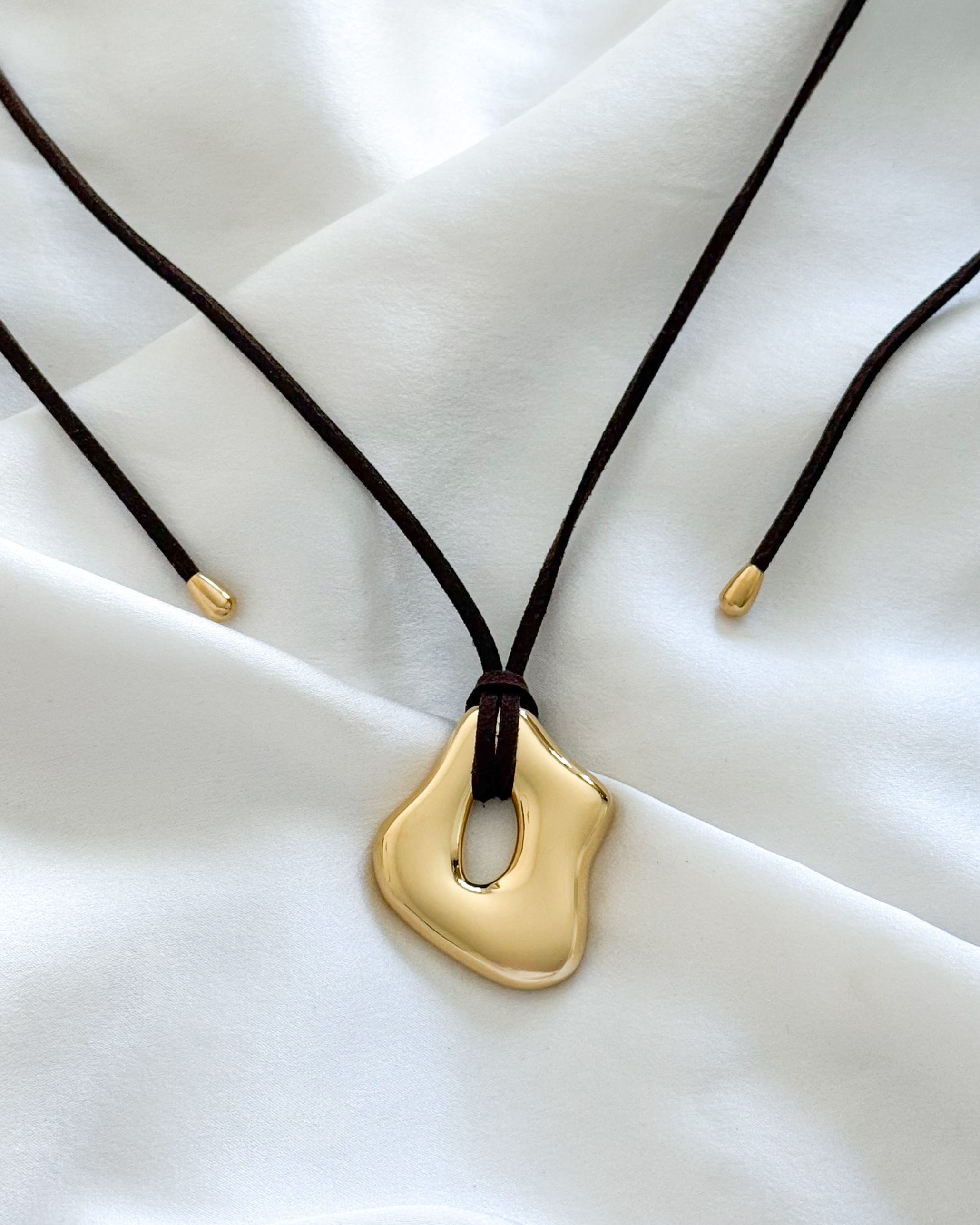 molten gold pendant necklace on a dark brown suede cord against a white fabric background