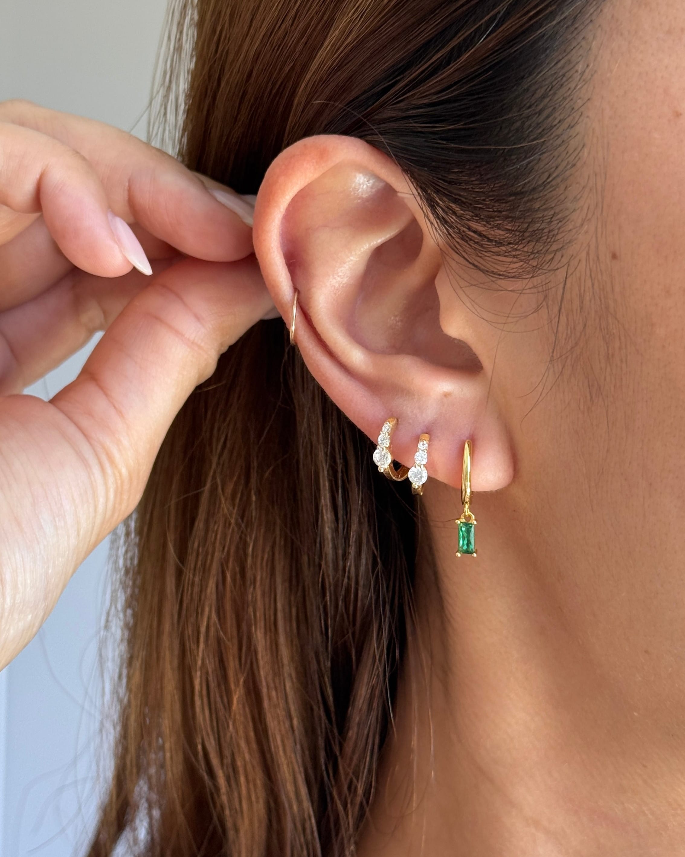 Close-up of a models ear wearing multiple earrings, including a small lumen gold hoop with a green rectangular stone and other delicate earrings, with a hand adjusting the hair.