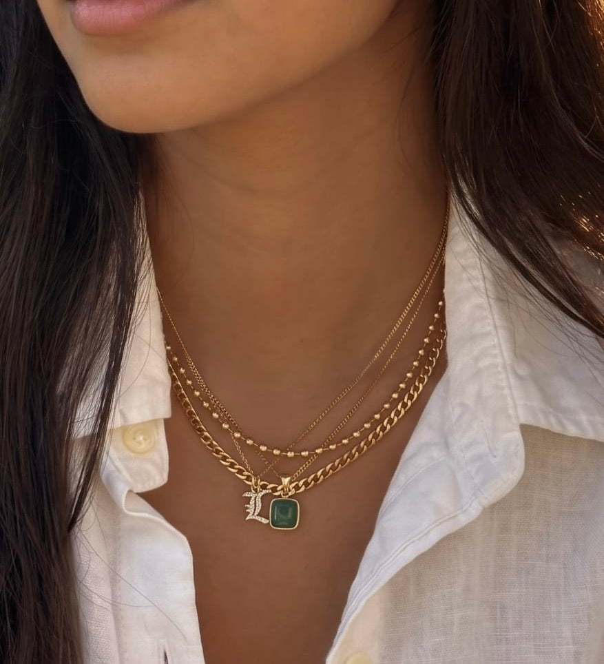 A close-up of a model wearing a layered gold necklace with a square green pendant and a gold initial 'L' charm, paired with a white linen button-down shirt.