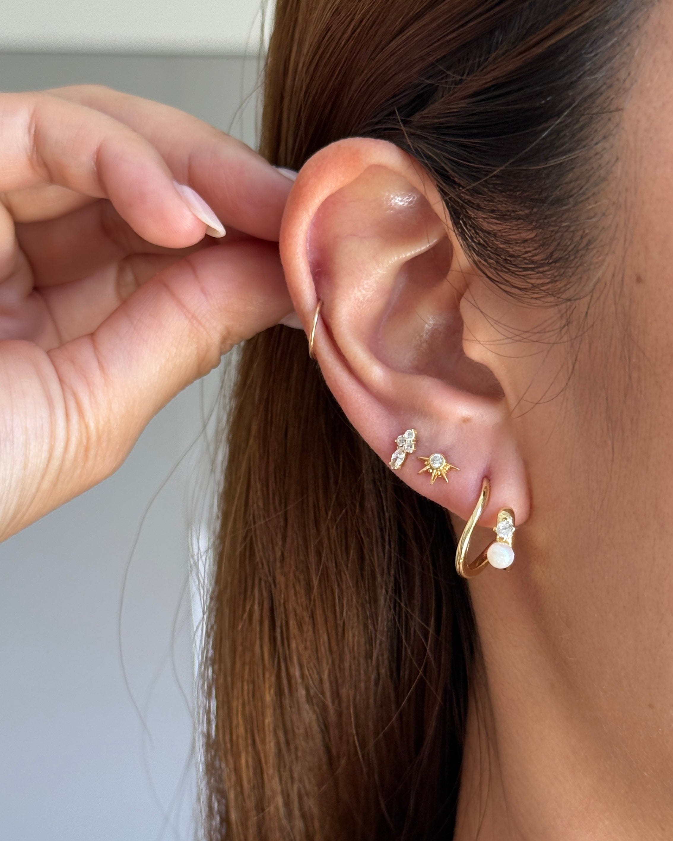 Close-up of a models ear wearing multiple small gold earrings, including a hoop, stud, and stacked bar earrings, with a hand adjusting the ear.
