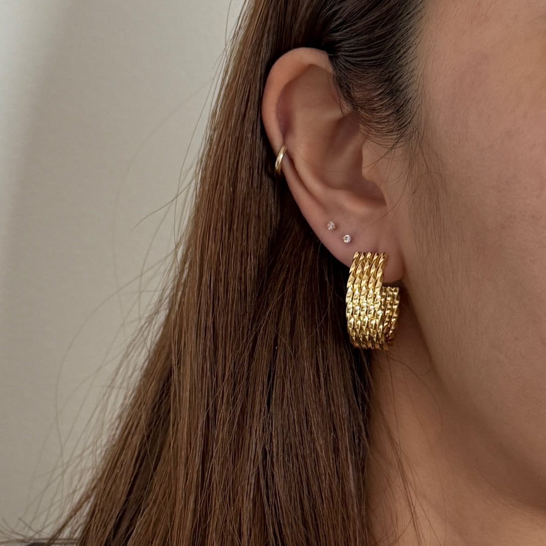Close-up of an ear wearing gold twisted hoop earrings with a neutral background