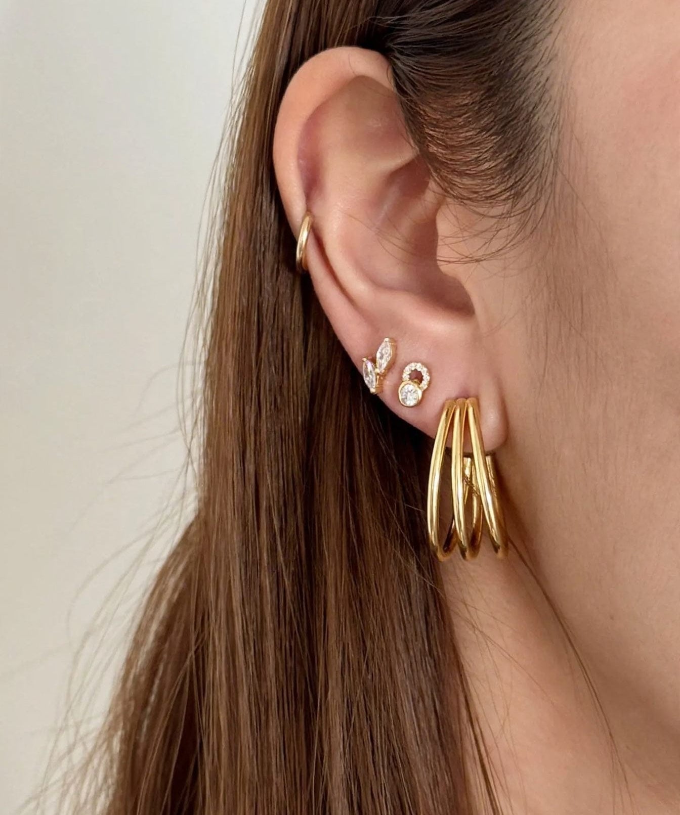 A close-up of a model's ear showcasing a stack of gold earrings, including a large twisted triple-hoop earring, a small diamond-accented hoop, and a delicate gold ear cuff, against a soft beige background.